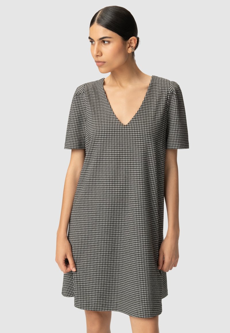 Kobieta z ciemnymi włosami w niskim kokie, ubrana w czarno-białą sukienkę w wzór hounds-tooth z dekoltem w serek i krótkim rękawem, patrząca w bok na jasnoszarym tle.