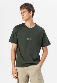 Dark green short-sleeve t-shirt made of cotton with a crew neckline and white printed logo on the chest, paired with light beige cargo pants.