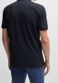 Polo noir à manches courtes, avec une texture subtile et des fentes latérales, associé à un pantalon bleu. Vue de derrière.