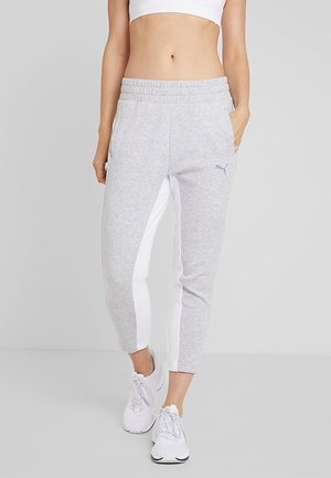 Kobieta ubrana w jasnoszare spodnie dresowe z białymi paskami po bokach, białe trampki i biały krótki top, stojąca na jednolitym tle.