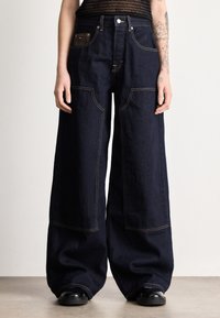 Άτομο που φοράει σκούρα τζιν με φαρδιά μπατζάκια και μεγάλες μπροστινές τσέπες, μαύρο τοπ και μαύρα παπούτσια, στέκεται σε φωτεινό πάτωμα μπροστά σε απλή λευκή τοίχο.