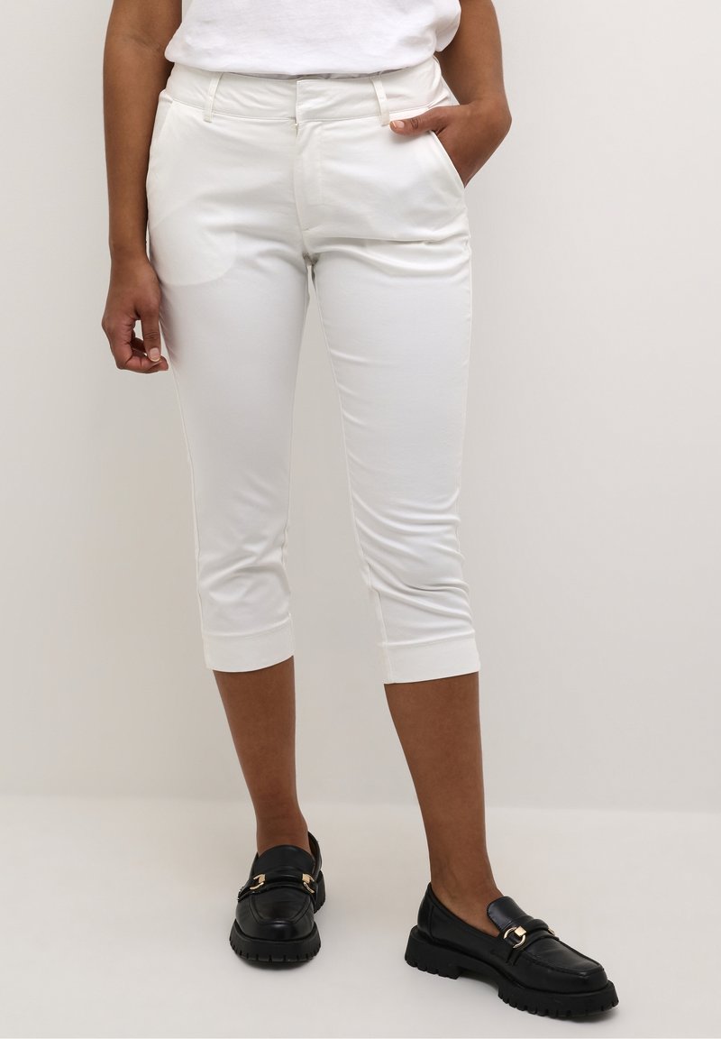 Femme portant un pantalon court blanc et des mocassins noirs sans lacets, debout, une main dans la poche, sur un fond uni.