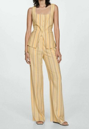 Woman wearing sleeveless yellow-beige vertical striped vest and matching wide-leg pants, standing against a plain light background.