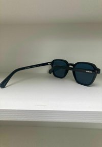 Zwarte rechthoekige zonnebril met donkere lenzen, kunststof frame en een recht armpatroon. Bevat merknaam en UV-bescherming categorie op de arm.