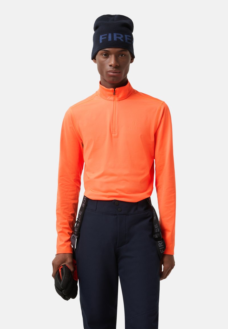 Pomarańczowa, wysokowidoczna bluzka z długim rękawem i zamkiem błyskawicznym, zestawiona z granatowymi spodniami z szelkami. Czarna czapka z napisem „FIRE” w niebieskim kolorze.
