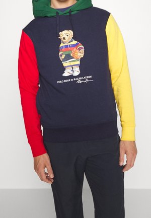 Person trägt einen Colorblock-Hoodie mit roten, gelben, grünen und blauen Bereichen, auf dessen Vorderseite ein Polo Bear mit einem Basketball abgebildet ist.