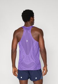 Lila Sporttop mit strukturiertem Mesh-Design, Racerback-Stil und einem kreisförmigen Detail in der Mitte. Getragen mit navyfarbigen Shorts.