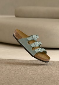 Πράσινο σανδάλι Birkenstock με τρεις ρυθμιζόμενες ασημένιες αγκράφες και φελλό σόλα, αιωρείται πάνω από μπεζ χαλί και φόντο.