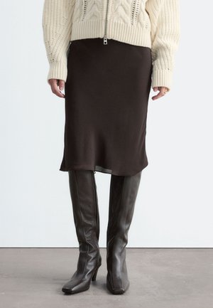 Femme portant un pull en maille crème, une jupe midi marron foncé et des bottes en cuir marron foncé à talons montant jusqu'au genou, debout sur un sol uni.