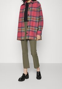 Plaid wool shirt jacket in pink with green and orange accents, button closures, worn over a black top and paired with olive green pants and black shoes.