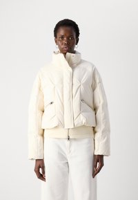 Persoon in een gewatteerde off-white pufferjack met ritszakken en bijpassende off-white broek, staand tegen een effen achtergrond.