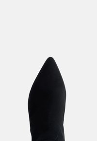 Pantofii negri din suede cu vârful ascuțit, pe un fundal alb simplu, văzuți dintr-o parte, evidențiind zona vârfului pantofului.