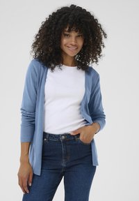 Cardigan aperto di un azzurro chiaro realizzato in un tessuto morbido, dotato di maniche lunghe e vestibilità rilassata, abbinato a una maglietta bianca e jeans blu scuro.