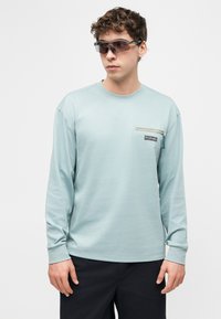 Νέος άνδρας που φοράει ανοιχτό μπλε πουκάμισο με μακρύ μανίκι και τσέπη με φερμουάρ στο στήθος, μαύρο παντελόνι, κοιτώντας προς τα πλάγια, φοράει σκουρόχρωμα γυαλιά ηλίου.