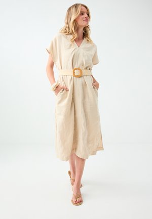 Femme blonde en robe midi beige avec une large ceinture et des poches, portant des sandales et des bracelets beiges, debout sur un fond blanc.