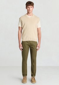 Beiger ärmelloser Pullover über einem kurzärmeligen cremefarbenen Hemd, kombiniert mit olivgrünen Hosen und braunen Wildlederschuhen.