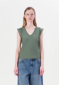 Olivengrønn tanktopp med V-hals, med blondebesatt skulderkapper. Kombinert med lyseblå, løstsittende denimjeans.