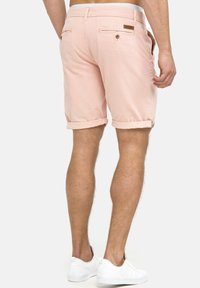 Shorts en coton rose avec ourlets roulés, poches arrière et étiquette en cuir. Portés avec des baskets blanches. Texture lisse et coupe ajustée.