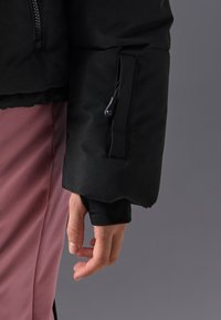 Mâneca unei jachete negre căptușite cu un buzunar cu fermoar. Textură netedă cu manșete elastice. Mâna vizibilă este relaxată, evidențiind potrivirea articolului vestimentar.