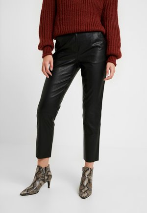 Osoba w czarnych skórzanych spodniach, bordowym dzianinowym swetrze i botkach w wzór skóry węża stoi na jednolitym białym tle.