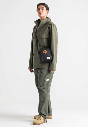 Giacca verde oliva con due tasche frontali, abbinata a pantaloni coordinati. Borsa a tracolla nera con un accento a rombo marrone e zip. Scarpe beige.