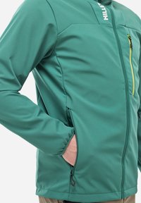 Türkise Softshell-Jacke mit Reißverschluss vorne, kontrastierendem gelben Reißverschluss-Anhänger und Seitentaschen. Verfügt über eine taillierte Passform und glatte Textur.