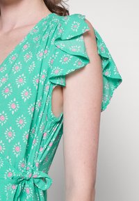Gros plan d'une personne portant une robe verte fleurie sans manches avec des détails à volants aux épaules et une ceinture nouée à la taille.