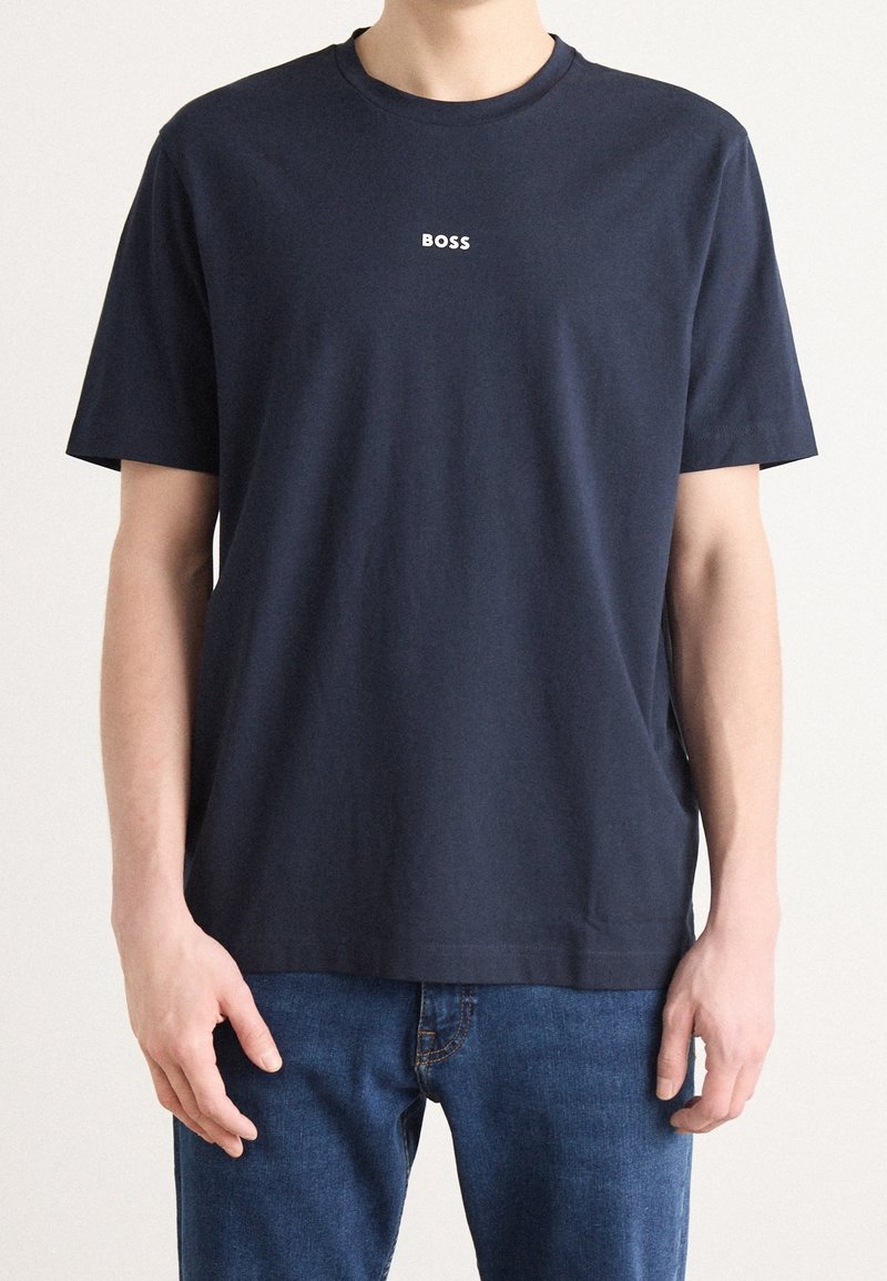 Personne portant un t-shirt bleu marine foncé uni à manches courtes avec le mot "BOSS" imprimé en petites lettres blanches sur la poitrine.
