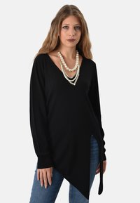 Tunic nera a maniche lunghe con scollatura a V e orlo asimmetrico. Abbinata a jeans, accentuata da collane di perle sovrapposte. Texture liscia.