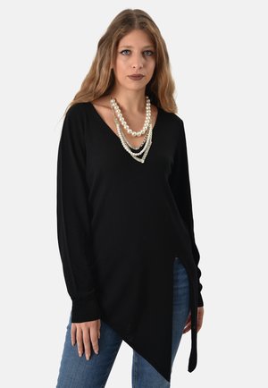 Tunic nera a maniche lunghe con scollatura a V e orlo asimmetrico. Abbinata a jeans, accentuata da collane di perle sovrapposte. Texture liscia.