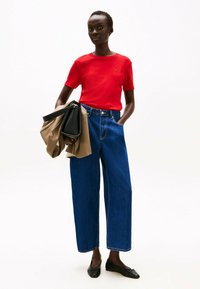 Rode shirt met korte mouwen, hoog getailleerde brede blauwe jeans, zwarte flats, vasthoudend een beige jas en een zwarte handtas. Glad materiaal en casual pasvorm.