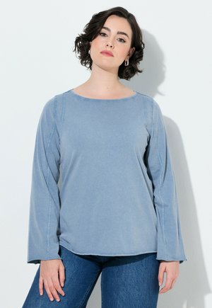 Frau mit kurzen dunklen Haaren, die ein hellblaues Langarmshirt und blaue Jeans trägt und an einer weißen Wand steht.