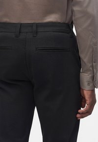 Vue de dos d’une personne portant un pantalon noir à poches passepoilées et une chemise beige à manches longues, la main droite détendue le long du corps.