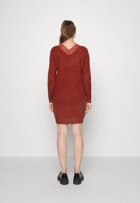 Vestito lavorato a maglia color ruggine con maniche lunghe, forma attillata e dettaglio a V sul retro con pizzo. Indossato con scarpe nere con suola spessa.