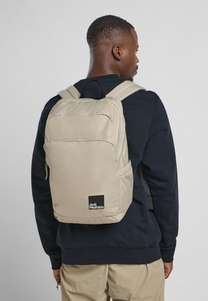 Beige backpack with padded straps, front zip pocket, and side mesh pocket. Features black logo label on bottom front. Smooth fabric texture.