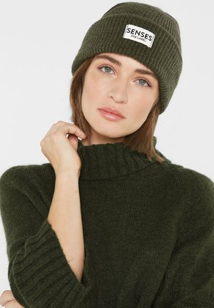 Dark green knit beanie with ribbed texture and a front patch labeled "SENSES .THE LABEL." Worn with a matching green oversized sweater.