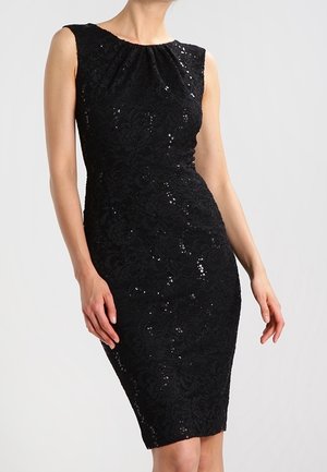 Femme portant une robe noire sans manches, longueur genou, avec de la dentelle et des détails subtils en sequins, debout devant un fond gris clair.