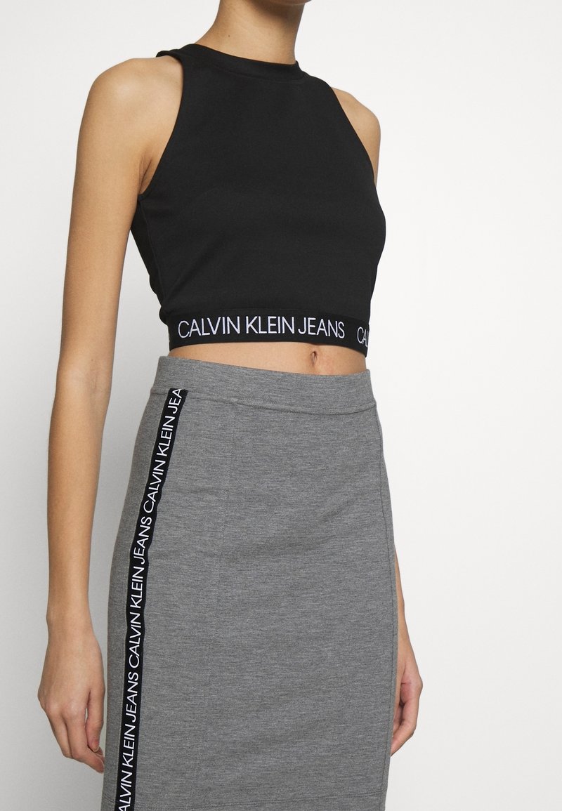 Kvinna iklädd en svart kortärmad tanktopp och grå kjol, båda med "Calvin Klein Jeans"-logoband i svart och vitt.