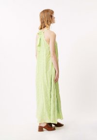 Hellgrünes, ärmelloses Maxikleid mit einer Rückenschleife, das ein strukturiertes Karomuster aufweist. Kombiniert mit braunen, hohen Sandalen.