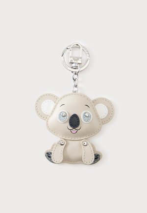 Porte-clés avec un design de koala beige souriant, yeux argentés, nez noir et détails cousus sur les oreilles et les pattes, attaché à un anneau en métal et une pince.