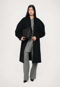 Caban negru din lână cu mâneci supradimensionate, top tricotat gri și pantaloni cu carouri gri. Ține în mână o geantă din piele neagră. Pantofi cu toc ascuțit negri.