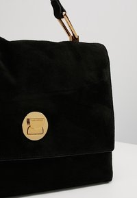 Zwarte suède handtas met een gouden ronde sluiting en hardware. Kenmerkt zich door een strak ontwerp en een gladde textuur. Compacte en gestructureerde vorm.