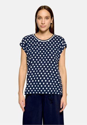 Femme de face portant une chemise à manches courtes bleu marine à pois blancs et un pantalon ample bleu marine sur un fond uni.
