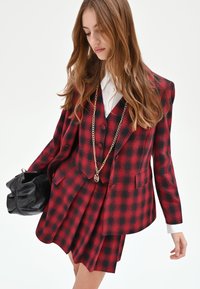 Completo a tre pezzi a quadri rossi e neri con blazer doppiopetto, gilet aderente e gonna plissettata, abbinato a una borsa nera con catena dorata.