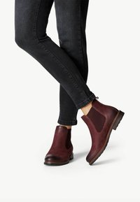 Burgundy nahkasaappaat nilkkamittaisilla kuminauha sivupaneeleilla, teksturoidulla pinnalla, pyöreällä kärjellä ja ruskeilla kumipohjilla. Yhdistetty mustiin skinny-farkkuihin.
