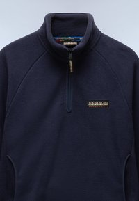 Marineblauer Fleece-Pullover mit hohem Kragen, halber Reißverschluss vorne und seitlichen Taschen. Mit Logo-Patch auf der linken Brust. Weiche Textur.