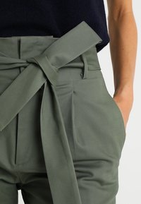 Gröna högmidjade byxor med ett brett tygbälte. Har sidofickor och en mjuk textur. Midjan har en rosett som detalj för knäppning.