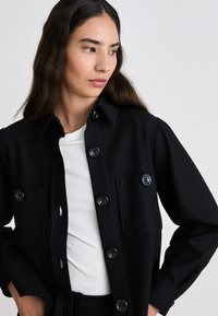 Camisa chaqueta de tela negra con botones, dos bolsillos en el pecho y un cuello de camisa. Presenta una textura suave y un corte ajustado.
