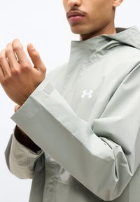 Άτομο που ρυθμίζει το μανίκι ενός ανοιχτόχρωμου γκρι μπουφάν Under Armour με λουράκι βέλκρο σε απλό λευκό φόντο.