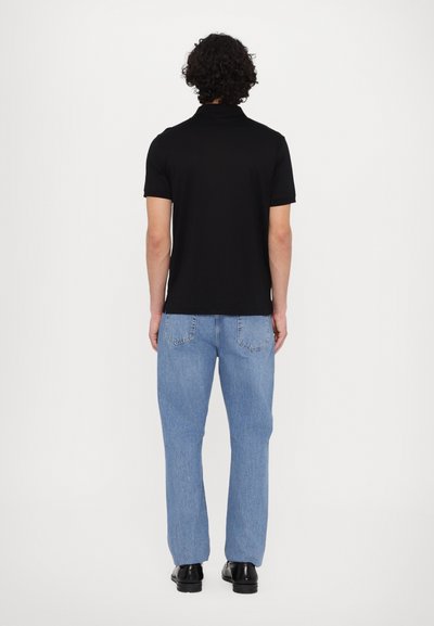 Μαύρο πόλο μπλουζάκι με κοντά μανίκια, συνδυασμένο με ανοιχτό μπλε τζιν παντελόνι και μαύρα παπούτσια. Ο ιστός φαίνεται λείος και εφαρμοστός.